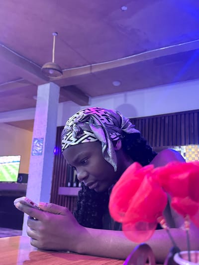 The image depicts a young girl with dark skin and braided hair, wearing a grey and white patterned headscarf, engrossed in her mobile phone. She is seated at a wooden table, with her hands holding the phone as she looks down at the screen. In the foreground, out of focus, are two vibrant pink flowers. The background suggests an indoor setting, possibly a restaurant or lounge, with ambient lighting casting purple and blue hues. A television screen displaying a sports event is visible in the left background, and a hanging light fixture is present on the ceiling. A light-colored pillar stands to the left of the girl. The image appears to be taken in Laminga, Nigeria, as indicated by the provided context.