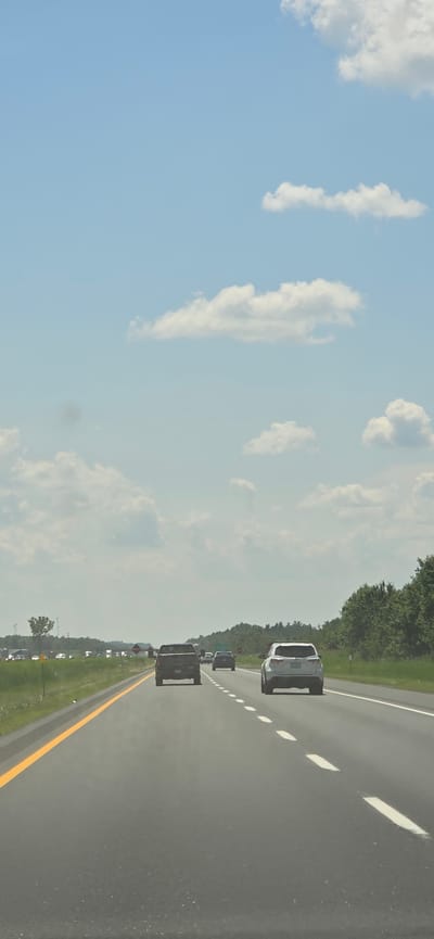 This is a vertical, eye-level, medium shot photograph taken from inside a vehicle traveling on a highway on a clear, sunny day. The highway stretches forward into the distance and is lined with green trees and vegetation on both sides. In the foreground, the asphalt road has yellow and white lines marking the lanes. Several vehicles are visible on the highway, all traveling in the same direction. The closest vehicles are a dark-colored pickup truck in the left lane and a white SUV in the right lane. In the distance, there are more cars and trucks, as well as some utility poles and trees. The sky is a bright blue with scattered white clouds. The image captures a typical highway scene with a focus on the road and the vehicles.