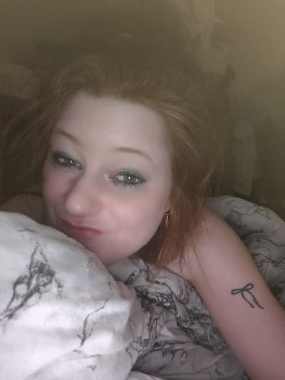 A selfie captures a red-haired woman in bed, partially covered by a marble-patterned blanket. Her face is close to the camera, featuring green eyeshadow and a slight smile. Her hair is tousled, with some strands near her forehead. She has a small earring in her visible ear. On her upper arm, a simple line drawing of a bow tattoo is visible. The background appears dimly lit with undefined objects.