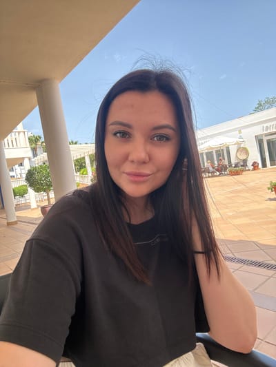 A young woman with long, dark hair and green eyes poses for a selfie outdoors. She is wearing a black t-shirt. Her right arm is bent and rests on a railing. The background shows a courtyard with tiled flooring, a white building with umbrellas and tables outside, and architectural elements like columns and balconies. The location is identified as Foia L'om, Spain. The sky is clear and blue.