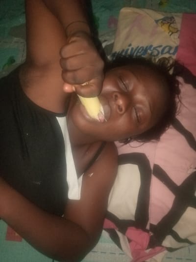 A woman with dark skin lies on a patterned bed with closed eyes. She wears a black tank top with a white stripe. She holds a peeled banana up to her mouth with her fist, seemingly eating it. Her hair is dark, and she wears small hoop earrings. The bedding has various patterns, including pink, white, and black designs.