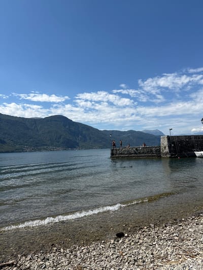 The image captures a sunny daytime scene on the shore of Lake Como in Abbadia Lariana, Italy. The foreground features a pebbled beach with a small wave breaking on the immediate shoreline. The lake water is calm with noticeable ripples, appearing clear near the shore and darker blue-green further out.
In the mid-ground, a dark stone pier or breakwater extends into the lake from the right side of the frame. It is constructed of large, dark grey stones and has a flat surface. Five individuals are present around this structure. On the pier, two individuals stand near the edge, one appearing to be preparing to enter the water. A third person is seated further back on the pier. Two additional individuals are visible in the water, partially submerged, swimming or floating near the pier.
To the right of the pier, a small white boat with a pointed bow is moored to the stone structure. A dark pole or light fixture is visible on the far right end of the pier.
The background is dominated by steep, forested mountains rising directly from the far side of the lake, extending across the entire horizon. A small village or settlement is discernible at the base of the mountains on the distant shore. The sky above is clear and bright blue with a band of scattered white cumulus clouds across the middle.