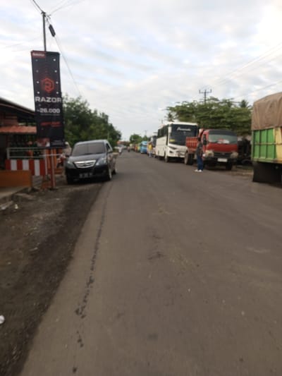 The image shows a street scene in Tocina, Indonesia. A black car is parked on the right side of the road, facing the camera. Further down the road, there are several buses and trucks parked in a line. A man in a grey shirt and blue pants is standing next to a red truck, looking towards the camera. A tall, black banner with red text advertising "RAZOR" is visible on the left side of the image. The sky is overcast with clouds. The environment appears to be a somewhat busy road with commercial establishments on the left.