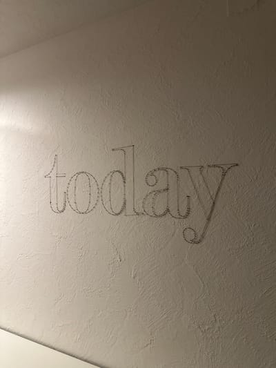 The image shows the word "today" formed by thin, dark wire sculpture attached to a textured, off-white wall. The lighting casts faint shadows on the wall, highlighting the uneven surface. The scene appears to be indoors, with a portion of a white surface visible at the bottom left, suggesting a floor or furniture. The wire sculpture is the sole focal point, crafted in a stylized font. The specific location in St. Märgen, Germany, is not discernible from the visual information provided in the image.