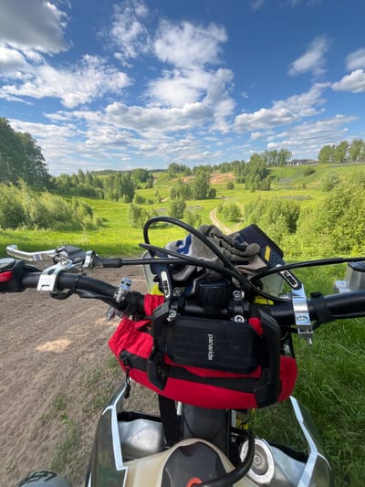 This is a first-person perspective shot from a motorcycle, stationary on a sandy dirt track, looking forward onto a rural landscape in Новочувашский, Russia. The scene depicts a bright, sunny day with a clear blue sky and numerous scattered white cumulus clouds.

In the immediate foreground, the handlebars of a dirt bike or enduro motorcycle are visible. The black handlebars feature silver clutch and brake lever assemblies on the left and right, respectively. A red button, possibly a kill switch, is on the left grip. A pair of grey, white, and blue motorcycle gloves, branded "100%", rests on the tank/handlebars. Centrally mounted on the handlebars is a black electronic device or holder, labeled "panaride", positioned above a red zippered tank bag or pouch. Portions of the motorcycle's white front fender, light-colored fuel tank, and black radiator shroud are also visible. The ground directly beneath the motorcycle is composed of light-brown sandy dirt with sparse green vegetation.

The midground features a winding dirt path or track that extends into the distance, flanked by lush green grass and numerous deciduous trees, suggesting a verdant, rolling terrain. This landscape consists of gentle hills and valleys covered in abundant greenery.

In the background, the rolling hills continue, densely covered with trees. On a distant hillside to the right, several modern houses or buildings with dark roofs are visible, indicating human settlement within the natural environment. The overall impression is one of a scenic rural area conducive to off-road motorcycling.