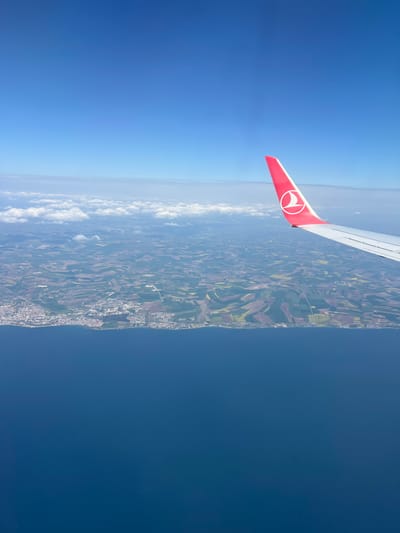 This aerial photograph, taken from an airplane window, captures an expansive daytime view over a coastal landscape. The scene unfolds beneath the aircraft's wing, which is prominently visible on the left side of the frame. The wing is silver-grey, with its red wingtip clearly displaying the white stylized bird's head logo of Turkish Airlines.

Below the wing, a diverse landmass stretches into the distance, characterized by a patchwork of agricultural fields in various shades of green, brown, and yellow. Scattered within this terrain are clusters of buildings and discernible roads, indicating human settlements and infrastructure. A distinct coastline marks the boundary between the land and a vast body of deep blue water on the right side of the image, likely a sea or large lake, appearing calm under the clear sky.

Above the land and water, the sky transitions from a lighter blue near the horizon to a deeper blue overhead. A layer of fluffy white clouds is visible at a lower altitude than the plane, running along the horizon line above the landmass. The bright, clear conditions suggest a sunny day, likely during a flight over the Tekirdağ region of Turkey, overlooking the Sea of Marmara.