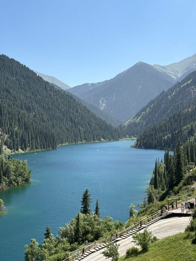 This media file captures a serene natural landscape with a crystal-clear blue lake and majestic mountains covered in lush green pine forests. The scene appears to be set in a mountainous region, possibly during the daytime on a clear, sunny day, judging by the bright sky and the sharp shadows cast by the trees and structures.
The lake itself is a prominent feature, with small white objects that could be boats or kayaks dotting its surface. Along the right side of the lake, a raised wooden boardwalk or viewing platform runs parallel to the shore. This platform is populated by several people, some walking and others observing the scenery. The presence of the boardwalk suggests a developed tourist area or a park that allows visitors to access and enjoy the natural beauty.
The mountains rise steeply from the edge of the forest, their peaks partially obscured by a light haze, hinting at their immense scale. The dense pine forest covers the lower slopes of the mountains, creating a striking contrast with the vibrant blue of the lake. The trees are a deep, rich green, indicating a healthy and thriving ecosystem.
In the foreground, closer to the viewing platform, there are more trees and vegetation, adding depth and texture to the scene. The overall impression is one of tranquility and awe-inspiring natural beauty. The people on the boardwalk seem to be enjoying a leisurely stroll or taking in the breathtaking views, contributing to the peaceful atmosphere of the location. There is no visible text in the image.