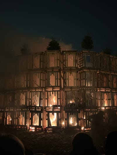 The image displays a large, towering bonfire at night. The photograph itself is oriented 90 degrees clockwise, making the tall structure appear to be lying on its side. When viewed in its natural upright orientation, the structure is a massive stack of wooden crates or pallets, forming a multi-level grid pattern.

The bonfire is actively burning, with bright orange and yellow flames emanating from within many of the individual compartments formed by the stacked wood. Sparks and burning embers are visible amidst the intense heat. On the upper left side of the burning structure, several small evergreen trees or branches are perched on top of some of the crates, seemingly integrated into the design of the bonfire. Wisps of smoke are rising from the structure, particularly visible against the dark sky on the left.

The setting is clearly nighttime, with a dark, deep blue-black sky dominating the background. The fire is the primary source of illumination, casting a warm, flickering orange glow that highlights the wooden structure and the rising smoke. In the lower right foreground, the silhouetted heads and shoulders of what appear to be spectators are visible, suggesting people are gathered to watch the event. The scene conveys a sense of warmth, light, and spectacle against the dark expanse of the night. No text is visible in the image.