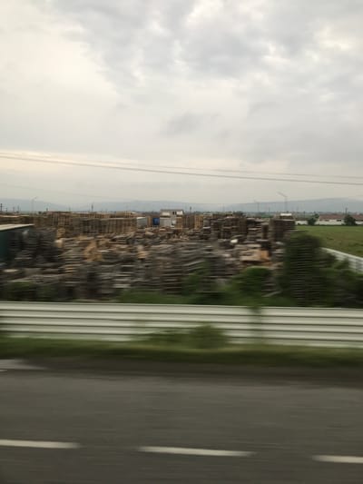 The image depicts an exterior scene, likely captured from a moving vehicle, showcasing an extensive outdoor storage area for wooden pallets and lumber, situated in what is identified as Nicolae Bălcescu, Romania. The sky is overcast with uniform grey clouds, indicating a cloudy day.

In the immediate foreground, a dark asphalt road with a faint white dashed line is visible on the left, exhibiting significant horizontal motion blur. Above the road, a multi-paneled metal guardrail, light in color, runs horizontally and is also heavily blurred. A strip of green vegetation, similarly blurred, separates the guardrail from the midground.

The midground is dominated by large, dense stacks of wooden pallets and various pieces of wood and lumber, occupying a wide expanse. Several structures are interspersed among the wood piles; these include what appears to be a partially constructed building made of concrete blocks, other industrial-type buildings, and a blue object or container.

In the background, beyond the storage facility, open fields with scattered residential or agricultural buildings are visible. Further in the distance, indistinct hills or mountains rise under the overcast sky. Multiple power lines stretch horizontally across the frame in the upper midground and background. Several tall, slender structures, consistent with wind turbines, are discernible on the distant horizon.

The entire image, particularly the foreground and parts of the midground, displays pronounced horizontal motion blur, indicating the photograph was taken while the camera was in motion. No people are visibly present in the scene.