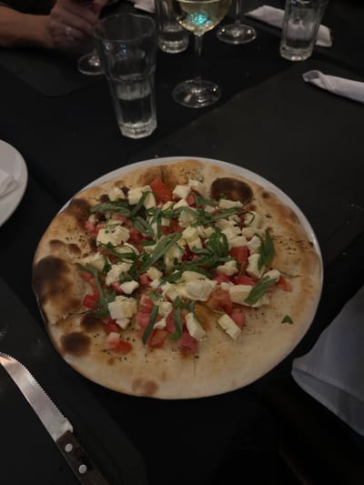 The image shows a pizza topped with tomato, mozzarella, and basil on a white plate. The pizza is on a black table. A knife is lying to the left of the pizza, while three empty glasses and a glass of white wine sit to the right of the plate. The image was taken in a restaurant in Santa Ana, Costa Rica, at the coordinates 9.935 latitude and -84.189 longitude.