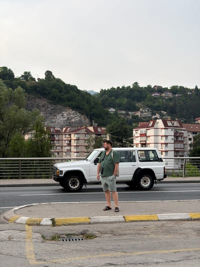 A man stands on a sidewalk in Konjic, Bosnia and Herzegovina, near a white SUV. The man is wearing a green short-sleeved shirt, light green shorts, and black sandals, and has a bag slung over his shoulder. The SUV is parked on a road that runs parallel to a bridge railing, with apartment buildings and a hillside covered in trees and houses visible in the background. The sky is overcast.