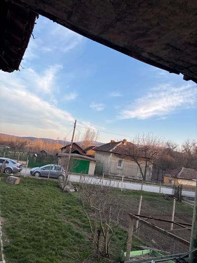 The image depicts an elevated, static view of a residential street in Erden, Bulgaria. The upper frame is defined by the dark, weathered underside of an architectural overhang, likely an eave, constructed of brick and wood. The sky displays a clear blue expanse with scattered, thin white clouds.

The immediate foreground features a downward-sloping grassy area, transitioning to bare earth containing small, defoliated shrubs and a rudimentary fence line composed of wooden posts and wire. No individuals are visible.

The midground is bisected by a road, on the left side of which two passenger vehicles are parked: a silver four-door sedan and a grey hatchback, both facing left. A wooden utility pole supporting multiple wires is positioned roadside. Across the road, several single-story houses with reddish-brown tiled roofs are present. One house is notable for a bright green garage door and structural damage to its roof. Another, a light-colored dwelling, displays one visible window. Bare deciduous trees are interspersed among the structures and property lines.

The background shows distant, undulating hills with sparse vegetation, illuminated by a low-angle light source suggesting either dawn or dusk.