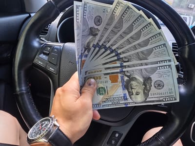 A person's hand, wearing a wristwatch, holds a fan of US $100 bills in front of a car's steering wheel. The steering wheel has integrated controls for volume, mute, and mode. The scene appears to be inside a vehicle, possibly in Eumseong-eup, South Korea, though the specific location is not confirmed by visual cues in the image. The lighting suggests it is daytime.