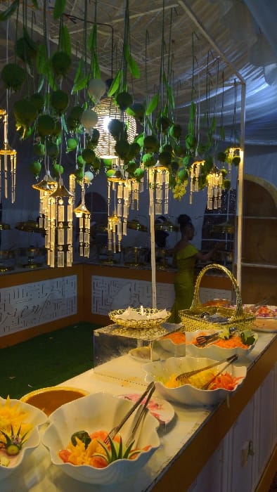 A banquet table is set with ornate white plates filled with a colorful salad. Silver cutlery rests on the plates. A gold basket sits prominently, and intricate gold decorations adorn the space. Behind the table, a woman in a green dress stands near what appears to be an elaborate floral and chandelier arrangement with artificial flowers and glass pendants.
