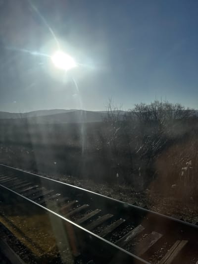 The image captures an outdoor scene near Bumbăta, Moldova, depicting a set of railroad tracks extending diagonally from the lower-left to the center-right. The tracks consist of two parallel steel rails secured by wooden sleepers embedded in a bed of gravel ballast. Strong sunlight from a bright, intensely flaring sun in the upper-left quadrant creates significant glare and reflections across the tracks and the foreground, suggesting the photograph was taken through a window. To the right of the tracks, the terrain consists of dark, possibly fallow ground with sparse, dry vegetation and several leafless deciduous trees or shrubs. In the midground, a series of rolling hills or low mountains are silhouetted against a hazy, bright sky, which transitions from pale blue near the sun to a clearer blue in the upper-right. No individuals, animals, or prominent structures other than the railway infrastructure are visible. The barren trees indicate a late autumn or winter season.