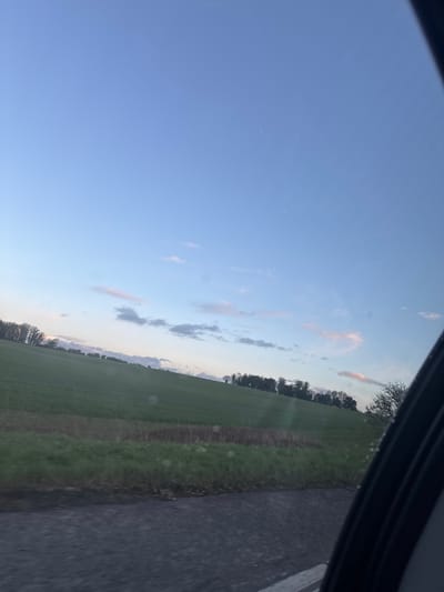 The image depicts a rural landscape within the Test Valley, United Kingdom, observed from a moving vehicle. The scene is dominated by a light blue sky with scattered cirrus and cumulus clouds, some exhibiting faint pink and orange tints indicative of low sun angle (dusk or dawn). A large, sloping green field occupies the midground, bordered by a strip of darker, possibly tilled earth, and a grassy verge in the immediate foreground. A line of deciduous trees forms a distant tree line along the field's horizon. A dark asphalt road, slightly blurred, is visible at the bottom, and the dark frame of a vehicle window or door partially obscures the far right edge of the image. Small, circular light reflections or imperfections are present on the glass surface. No individuals or specific actions are discernible.