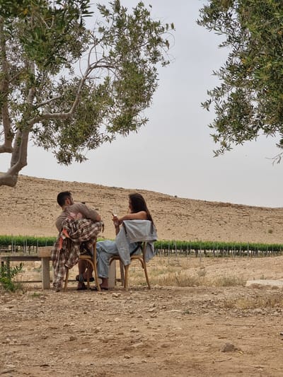 Two people are seated on chairs in an outdoor setting. One person holds a clear glass, possibly containing a beverage. The location appears to be rural, with an unpaved ground surface and a backdrop of a high, tan-colored wall or embankment. A row of green plants, likely bushes or shrubs, runs parallel to the wall. A tree with green leaves is visible to the left of the people. Given the city of Mizpe Ramon, Israel, the location is likely an arid landscape or an outdoor area near vineyards. The nature of their interaction is unclear, but their proximity suggests they are conversing or sharing a moment.