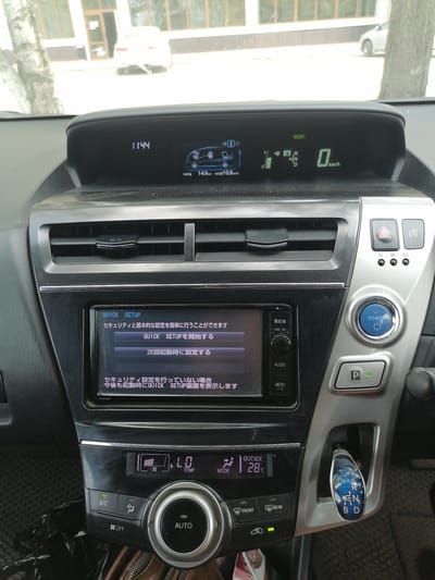 The image shows the interior of a car, specifically the dashboard and center console. A digital display in the upper part of the dashboard shows the time as "11:44" and other vehicle information including speed and average fuel consumption. Below this, a touchscreen display presents menu options in Japanese, with one option highlighted. To the right of the touchscreen are various buttons, including a power button and a parking brake release. The center console features climate control buttons and a gear shifter. The car appears to be parked. The provided context states the location is Lesozavodsk, Russia.