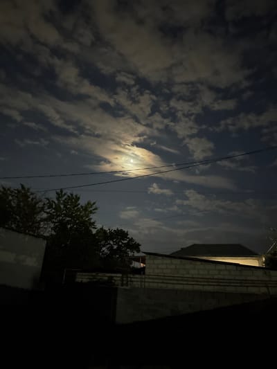 The image depicts a nighttime scene, oriented vertically, with a prominent moon visible through scattered clouds. The sky is a deep blue, with streaks of white and grey clouds partially obscuring the moon, which is bright and appears to be in its waxing or waning phase. A few faint stars are also visible.

In the foreground, there are several dark electrical wires stretching across the frame, suggesting an urban or suburban setting. To the right of the image, a building with a brick facade is visible. The building appears to be illuminated by artificial light, casting shadows and highlighting the texture of the bricks. A portion of the building's roof is also visible, covered with angled tiles. Adjacent to the building, there is a dense cluster of green foliage, likely a tree, silhouetted against the moonlit sky.

The overall mood of the image is serene and atmospheric, characteristic of a peaceful night. The time of day is clearly night due to the presence of the moon and the darkness of the scene. The weather appears to be partly cloudy, with the clouds allowing glimpses of the moon. The combination of the moon, clouds, building, and tree suggests a location in a settled area, possibly residential or semi-industrial, in Nogir, Russia. There is no visible text in the image.