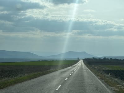 The image displays a two-lane paved road, marked with double broken white lines, extending straight into the distance under a partly cloudy sky in the vicinity of Zvegor, Bulgaria. The road surface appears damp or reflective, particularly where a prominent sunbeam or lens flare originates from the upper left and streaks diagonally across the sky and down to the mid-ground. To the left of the road are agricultural fields, some tilled and dark brown, others showing green vegetation. Distant, hazy blue-gray mountains form the horizon on the left. To the right of the road, similar fields are visible, alongside a dense, dark green line of trees or bushes running parallel to the roadway. The sky is predominantly covered by layered gray and white clouds. No people, vehicles, or animals are present.