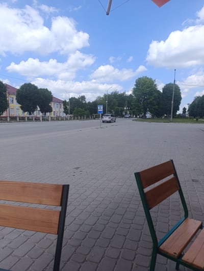 The image depicts a public square in Chemerivtsi, Ukraine, under a bright blue sky with scattered white clouds. In the foreground, two wooden benches with green metal frames are partially visible, suggesting a seating area. The ground is paved with gray cobblestones.
Beyond the seating area, a wide asphalt road stretches across the scene, with a few vehicles present. A silver SUV is parked on the left side of the road, near a blue sign featuring a white cross, indicating a medical facility or first aid point. Further down the road, other cars are visible in the distance.
To the left of the road, a long, multi-story building with a colorful facade (yellow, pink, and blue accents) and numerous windows is situated behind a decorative metal fence. Several mature trees with lush green foliage are planted in front of the building and along the street.
On the right side of the road, more trees and greenery are present, along with a utility pole and streetlights. The overall atmosphere is one of a quiet, daytime scene in a Ukrainian town.