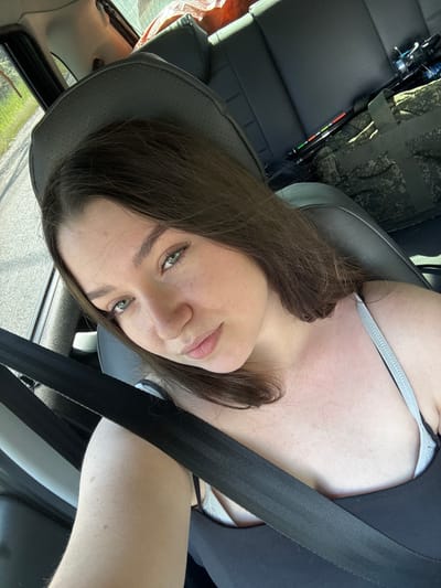 A young woman with long, dark hair and green eyes is seated in the passenger seat of a vehicle, looking at the camera. She is wearing a black tank top and a grey bra strap is visible. A black seatbelt crosses her chest. Fishing rods and a camouflage bag are visible in the background. The vehicle appears to be on a road with greenery on the left side. The image appears to have been taken in Pesterevo, Russia.