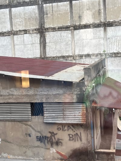 The image depicts a weathered building in Dumai City, Indonesia, characterized by its decaying facade and corrugated metal roofing. The background is dominated by a large, imposing concrete structure, possibly an abandoned industrial building, with visible signs of age and neglect, including cracks and stains. In the foreground, a smaller structure with a rusty red metal roof shows signs of disrepair, with exposed concrete walls and corrugated metal window coverings. Graffiti, including what appears to be "BIM" and "BIN" is visible on the concrete wall, along with other unreadable markings. The overall impression is one of urban decay and the passage of time, with nature beginning to reclaim the built environment, as evidenced by the small plants growing on the concrete structure.