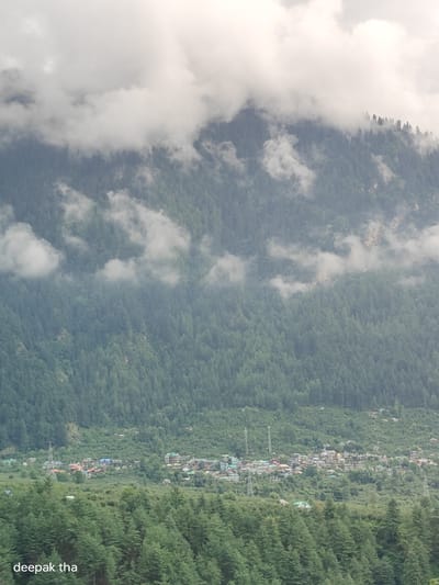 The image depicts a scenic mountainous landscape dominated by dense pine forests. The upper portion of the image is shrouded in thick, white clouds that partially obscure the mountain peaks. Below the clouds, the slopes are covered with an extensive, dark green forest. In the middle ground, a cluster of buildings forming a small town is visible, nestled amongst the trees. These buildings appear to be a mix of residential and possibly commercial structures, with some featuring colorful roofs. Telecommunication towers are also present in the vicinity of the town. The foreground is filled with more pine trees, creating a layered effect that leads the eye towards the distant town and mountains. The overall impression is one of a natural, serene environment with a human settlement integrated into the landscape. The text "deepak tha" is present in the lower left corner of the image, which appears to be a watermark. Based on the prompt's constraints, this image is not identifiable as being within the city of Baran, India.