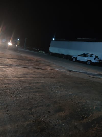 At night, in Semionovca, Moldova, a white Dacia Duster is parked on a ramp, likely near a truck trailer. The scene is dimly lit by artificial light sources, casting long shadows and highlighting the textured, worn surface of the asphalt. In the background, a large truck trailer is visible, its side illuminated by overhead lighting. To the left, another vehicle, possibly a car or van, is partially visible with its headlights on, creating a glare. The environment suggests an industrial or logistical area, possibly a loading dock or a transport hub, given the presence of the trailer and the ramp. The ground appears to be a rough asphalt surface with tire marks and some debris. The overall atmosphere is one of nocturnal activity with limited visibility.