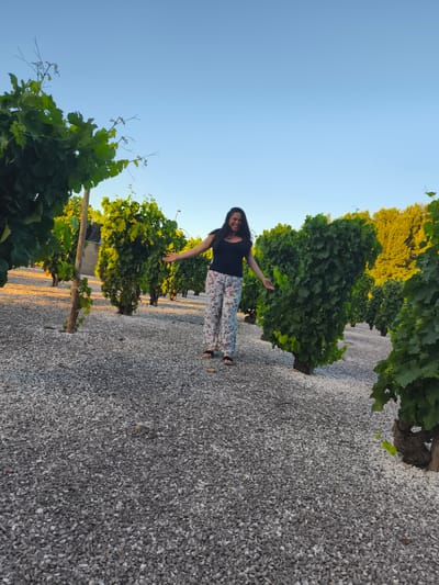 A woman stands in a vineyard in Haro, Spain, with rows of grapevines stretching into the distance under a clear blue sky. She is wearing a black top and patterned pants, with her arms outstretched. The ground is covered in white gravel, and the grapevines are lush and green, with some showing developing grapes. The sun is low in the sky, casting a warm glow on the scene.