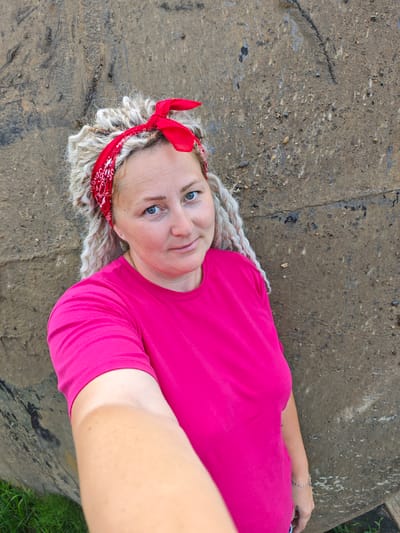 A woman with blonde dreadlocks and a red bandana tied around her head is taking a selfie in front of a concrete wall. She is wearing a bright pink t-shirt. Her left arm and hand are visible in the foreground as she holds the phone to take the picture. She has blue eyes and is looking at the camera with a slight smile. The background is a rough, textured concrete surface with some dark stains and small pebbles. The bottom left of the image shows a glimpse of green grass. The scene is outdoors, possibly in an urban or industrial area of Novoe Mart'yanovo, Russia.