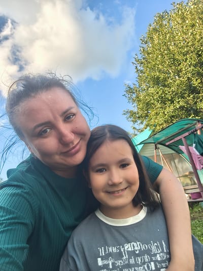 A selfie taken outdoors shows two people, an adult and a child, embracing and smiling. The adult, on the left, has dark hair pulled back and is wearing a dark green ribbed shirt. The child, on the right, has dark hair and is wearing a grey t-shirt with a graphic print. They are positioned in front of a blurred background that includes a green structure, possibly a greenhouse or canopy, and lush green foliage. The sky is visible above with scattered clouds. The scene suggests a casual, happy moment, likely in a residential or garden setting. It is highly probable this photograph was taken in Burayevo, Russia, given the context provided.