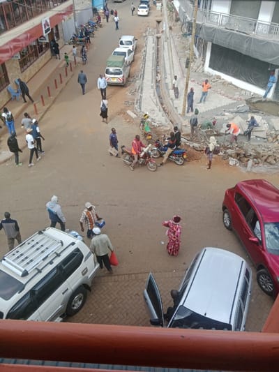 The image shows a street scene in Kapsabet, Kenya, at the intersection of Kisumu Road and Oloo Street. It is a busy street scene with many people walking and driving cars. Several people are walking on the sidewalk, some are crossing the street, and a few are standing around talking. There are many cars parked on the side of the street, and three vehicles in the middle of the road, one is a white SUV with a roof rack, one is a silver sedan, and one is a red compact SUV. Two motorcycles are being driven by men on the road. In the background, construction is underway on a new sidewalk. Workers are laying bricks and moving materials. The scene is a mix of urban life and construction. There is a lot of activity in the street and the atmosphere is bustling and energetic.