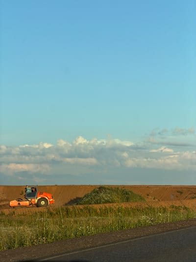 An orange steamroller is operating on a dirt road, compacting the soil. The road is surrounded by fields of dry grass and wildflowers. In the distance, there are rolling hills and a clear blue sky with scattered white clouds. The scene appears to be a rural landscape in the city of Derzhavino, Russia.