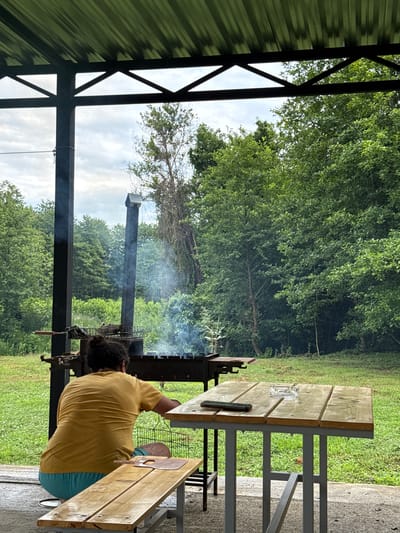 The image depicts a person tending to a barbecue pit under a covered structure, likely outdoors. The person, seen from the back, is wearing a yellow t-shirt and blue shorts and has dark, curly hair. They are positioned next to a grill that is producing smoke, suggesting food is being cooked. In the background, lush green trees and foliage create a natural setting. A picnic table made of light-colored wood with metal legs is visible on the right side of the frame, with an ashtray on its surface. The overall scene suggests a casual outdoor gathering or meal preparation. The lighting indicates it is daytime. The provided location context is Warcha, Abkhazia.