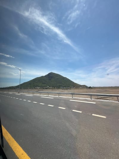 The image shows a road in Tuy Hoa, Vietnam, with a mountain in the background. The road is paved with asphalt and has white dashed lines marking the lanes. A yellow line is visible on the right side of the road. The sky is blue with some white clouds. The mountain is covered in green vegetation, and there appears to be a small structure or antenna on its peak. In the distance, there are some buildings and fields. A lamppost is visible on the left side of the road. The scene suggests a clear day, likely daytime, given the bright lighting. The image is taken from a low angle, possibly from a vehicle moving along the road. There is no visible text in the image.