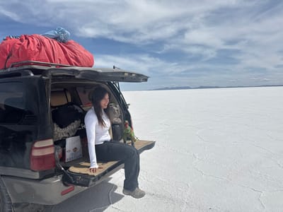 A single female individual is depicted seated on the open tailgate of a dark-colored SUV, parked on a vast, flat white salt plain, identified as Salar de Uyuni within Municipio Colcha K, Bolivia. The woman, appearing to be of East Asian descent with long dark hair, wears a white long-sleeved shirt, dark pants, and hiking shoes. She is positioned facing right, with her legs extending into the cargo area, and her head turned to gaze towards the distant horizon. Her expression is calm.
The SUV, likely a 4x4, has its rear hatch fully open and its tailgate lowered to form a seating surface. The vehicle's roof rack supports a large, bulky item wrapped in red tarp and a smaller item in blue. Inside the cargo area, various soft bags, a blanket, and a white box are visible. A small green dinosaur-like figurine rests on the tailgate surface to the woman's right.
The environment consists of an expansive white salt flat characterized by visible polygonal cracks on its surface, stretching to a distant horizon line where faint, low-lying mountains are discernable. The sky above is partly cloudy with patches of blue and white altocumulus clouds. The vehicle is stationary on the salt crust.