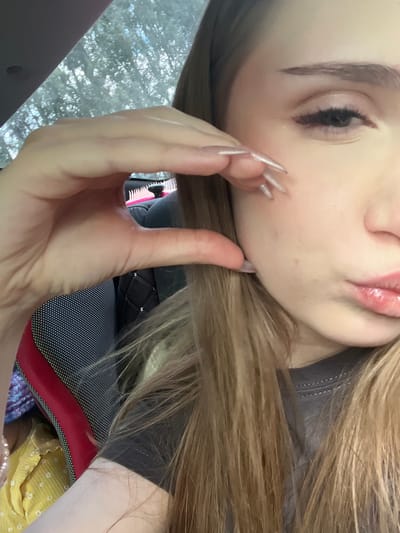 The image shows a close-up of a young woman's face and hand, taken from the chest up. She has blonde hair and is wearing a grey t-shirt. Her right hand is positioned near her cheek, with her fingers gently pinching her skin. Her left eye and part of her nose and lips are visible. The background suggests an indoor setting, possibly a car, with a headrest and a glimpse of foliage through a window. The lighting is natural, indicating it may be daytime. There is no indication of the city of Новая Отрадовка, Russia, in the image.