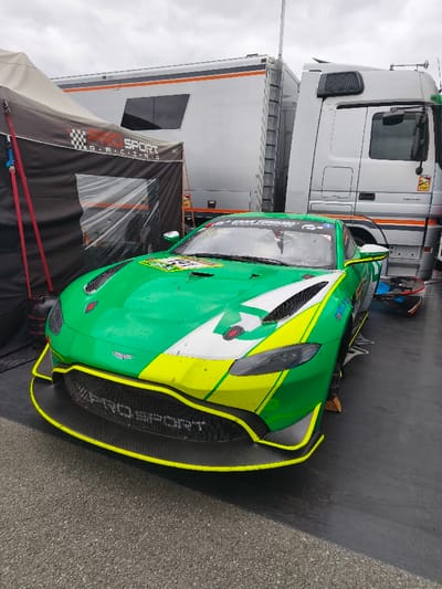 A green, white, and neon yellow Aston Martin Vantage GT3 race car is depicted stationary on an asphalt surface, likely within the paddock or pit area of the Sachsenring race circuit in Hohenstein-Ernstthal, Germany. The vehicle, elevated on visible supports, features "PRO SPORT" branding on its front grille and "GRAN TURISMO" and "FALKEN" decals. Its livery shows signs of track use with dirt and scuff marks. To the left, a black canopy tent with "PRO SPORT R-RACING" identification is partially erected. Behind the tent and to the right, a silver semi-trailer race transporter with orange stripes is parked. Adjacent to it on the right is the cab of a white Mercedes-Benz semi-trailer truck, also a race transporter, displaying "1830" on its side. The sky is overcast. No individuals are visible in the scene.
