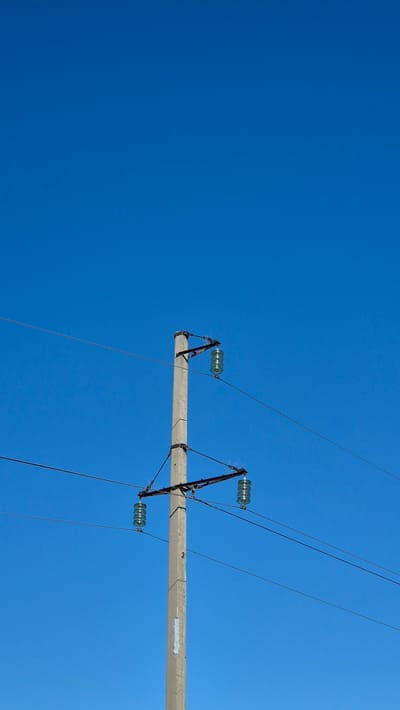 The image is a vertical, medium shot of a concrete utility pole against a clear, deep blue sky. The pole is positioned slightly left of center in the frame. It has two horizontal crossbars, each supporting a cluster of three clear glass insulators. Power lines are visible extending horizontally from the pole and disappearing out of frame to the left and right. The sky is uninterrupted by clouds or other objects. There are no people or animals visible in the image. The scene appears to be outdoors, possibly in an open area, as suggested by the expanse of sky and the presence of utility infrastructure. The overall impression is one of a clear, bright day.