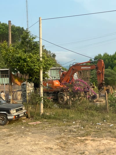 The image depicts an outdoor scene near the city of Myui, Vietnam. An orange Doosan excavator is parked partially off-road, adorned with purple flowers. The excavator is near some wire mesh. Power lines are visible overhead. A gray vehicle is parked nearby, with a person visible inside. The ground appears to be dirt and grass, with scattered debris. A stack of material is visible to the left of the excavator. The environment includes trees and foliage in the background.