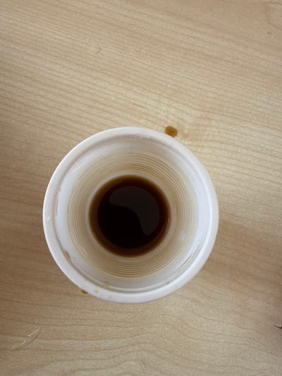 An overhead view displays a white disposable cup resting on a light-brown, horizontally striped wood-grain surface in Aragats, Armenia. The cup's interior walls exhibit concentric ridges and contain a shallow layer of dark brown liquid at the base, reflecting an overhead light source. Faint yellowish-brown residue is visible along the inner walls above the liquid level. An irregular dark brown liquid spill is present on the wood surface to the upper right of the cup, accompanied by smaller, diffuse liquid marks nearby. No individuals, actions, or interactions are depicted.