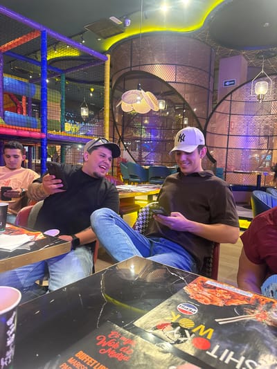 This is a vertical, medium shot photograph taken inside a restaurant. The primary subjects are three young men, two of whom are clearly visible, and a portion of a third. The man in the foreground, seated on the right, is wearing a black t-shirt and a baseball cap, and is laughing while holding a smartphone. He has tattoos on his arm. To his left, another man in a brown t-shirt and a white baseball cap is also smiling and holding a smartphone. Behind them, a third man in a light-colored t-shirt can be seen looking down at his phone. The setting appears to be a modern restaurant with elaborate decor, featuring metallic screens with intricate patterns and colorful ambient lighting. In the background, a brightly lit children's play area with blue, purple, and yellow netting is visible. A menu or placemat with text in Spanish, including "Et mej Wo Madria" and "BUFFET LIB," is spread across a table in the foreground. The overall mood seems casual and jovial, suggesting a social gathering, possibly a family outing or a meal with friends. The lighting indicates it is likely evening. The text on the menu suggests the restaurant is located in Madrid, Spain.