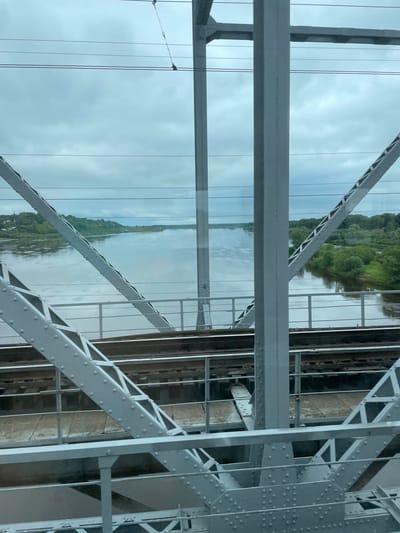 The image shows a view from a bridge over a river in Kotelnich, Russia. The bridge is made of metal girders and has railway tracks on it. The river is wide and calm, with green trees lining its banks. The sky is cloudy and overcast. There are no people or animals visible in the image. The scene appears to be peaceful and serene.