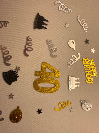 The image displays birthday-themed confetti scattered on a light-colored surface. A large glittery gold "40" is centrally positioned. Surrounding it are various shapes including stars, streamers in gold and silver, cake silhouettes in black and silver, and balloon cutouts. A "Happy Birthday" phrase is visible in gold at the top. The composition suggests a celebration for a 40th birthday.