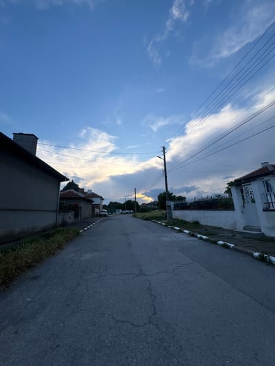 The image depicts a street scene in the city of Tenevo, Bulgaria. The perspective is from ground level, looking down a paved road lined with houses and vegetation. The sky is a mix of blue and grey clouds, with a hint of yellow light suggesting a sunset or sunrise. Power lines are visible, strung across the sky and supported by utility poles. The road itself is cracked and shows signs of wear. On the left side of the road, a dark-colored house with a chimney is prominent. On the right side, a white house with an ornate metal gate is visible. Several cars are parked along the street in the distance. The overall impression is that of a quiet, residential street in a rural or small-town setting.