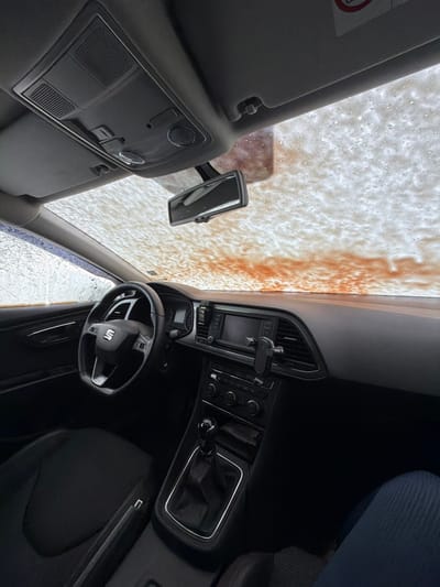The image displays the interior of a vehicle, consistent with a SEAT model, viewed from the front passenger side looking forward. The primary action visible is the vehicle undergoing a car wash, indicated by white foam and reddish-orange liquid partially covering the exterior of the windshield. The car's interior features a black dashboard with an infotainment screen centrally located. A black claw-style phone mount is attached to an air vent to the right of the screen. Below the screen are various climate and media controls. The driver's area includes a black multi-function steering wheel with the SEAT logo. A manual gear shifter, featuring a black knob and a chrome-trimmed base, is present on the center console. In the bottom right foreground, the lower legs of an individual wearing blue trousers are partially visible, suggesting a passenger is present. Overhead, a grey headliner contains an overhead console with controls, and a rearview mirror is affixed to the windshield. The precise location within Tserovo, Bulgaria, cannot be determined from the visual information provided in the image.