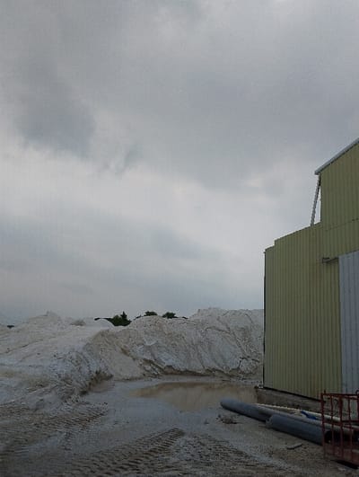 The image shows a large pile of white sand in the foreground. The sand is piled high, creating a large, rounded mound. There is a small puddle of water in the middle of the sand. On the right side of the image, a large yellow metal building stands partially visible. The building has a corrugated metal wall and a white roof. The sky is overcast with grey clouds. The image was taken in a remote area on the outskirts of Pangasia, Bangladesh (latitude: 22.693, longitude: 90.601). There are no people in the image. There are two large grey pipes lying in the foreground near the building, and other assorted metal debris in the foreground.