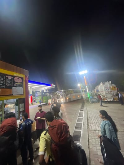 The image depicts an outdoor scene at night, likely a gas station and bus stop in Kajheri, India. In the foreground, several people are gathered around a food stall or counter. Some are holding backpacks, suggesting they are travelers. A man in a maroon t-shirt is holding a bowl. To the left, a yellow menu board is visible.

In the mid-ground, a green FlixBus is parked. Behind it, a white tanker truck with an "HP" logo is also present. Several other buses and vehicles are visible in the background, illuminated by bright overhead lights and the lights of the vehicles themselves. The environment appears to be paved, with drainage channels running along the side. The sky is dark, indicating it is nighttime. There are signs that say "NO PARKING." The overall atmosphere suggests a bustling transit point at night.
