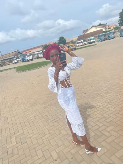 A woman stands on a brick-paved surface, holding a smartphone up to take a selfie. She's wearing a white lace dress, white heels, and a maroon headwrap. The background reveals several parked buses and buildings, suggesting an urban location. The scene is in Otta, Nigeria, as inferred from the buses. The sky is partially cloudy.